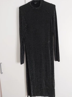 Glittrig svart långärmad blus - Snygg svart blus med glittereffekt och lång ärm. Modellen är rak och har hög hals, vilket ger en stilren look. Materialet är stretchigt och skimrar i ljuset, perfekt för att sticka ut lite extra.