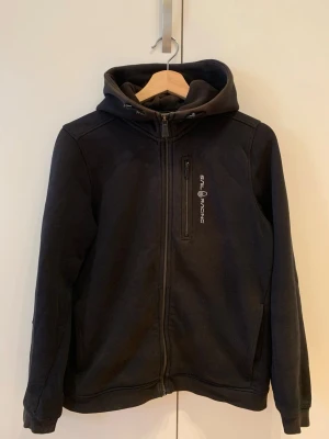 Svart hoodie från Sail Racing - Säljer en svart hoodie från Sail Racing med dragkedja framtill och bröstficka med logga. Hoodien har justerbar huva och två sidofickor. Tillverkad i mjukt material som känns skönt mot huden. 
