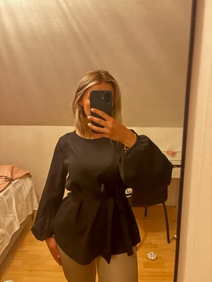Svart blus med knytband i midjan - Snygg svart blus med rund halsringning och ballongärmar. Blusen har ett brett knytband i midjan som ger en markerad siluett. Tillverkad i ett slätt tyg som ger en elegant känsla. Perfekt att styla med både jeans och kjol. den är en S men ganska oversized därav skriver jag att det är strl M. Helt felfri!