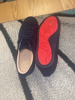 Mörkblå sneakers med röd sula - Snygga mörkblå sneakers i mocka med klassisk låg profil och ikonisk röd sula. Skorna har matchande mörkblå snören och diskret broderad text på hälen. Insidan är ljusbeige och ger en lyxig känsla. Perfekta för dig som vill sticka ut med stilrena detaljer.