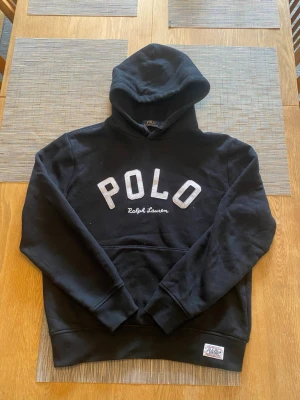 Svart hoodie från Polo Ralph Lauren - Säljer en svart hoodie från Polo Ralph Lauren med stor POLO-text i vitt på bröstet och broderad Ralph Lauren-logga under. Klassisk känguruficka framtill och huva. Mjuk bomullsblandning och ribbade muddar vid ärmar och nederkant.