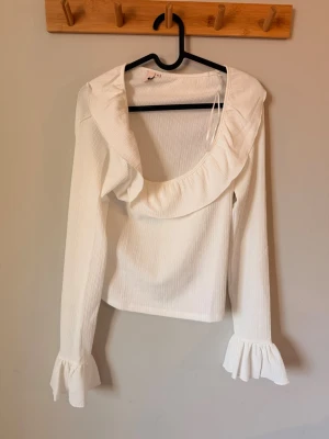 Vit blus med volangkrage och volangärmar - Supersöt vit blus med lång ärm, stor volangkrage och volangdetaljer vid ärmsluten. Blusen har en lätt ribbad struktur och en feminin, romantisk look. Perfekt att styla med jeans eller kjol för en trendig outfit. Från Nelly. Lapparna finns kvar!