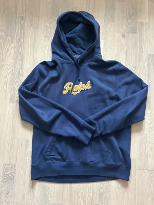 Mörkblå hoodie från Polo Ralph Lauren - Snygg mörkblå hoodie från Polo Ralph Lauren med broderad gul text 'Ralph' på bröstet. Klassisk huva med dragsko och känguruficka framtill. Perfekt för en avslappnad och trendig stil. Med inga fläckar eller märke