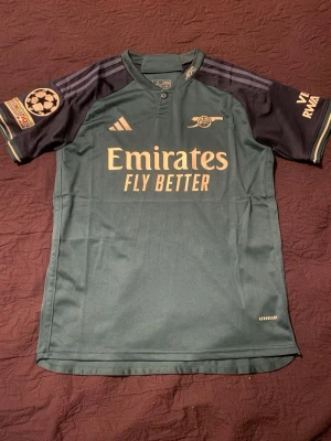 Arsenal fotbollströja Martinelli 11 - Mörkgrön Arsenal bortatröja från Adidas med Martinelli och nummer 11 på ryggen. Tröjan har svarta ärmar, Champions League-märke på ena ärmen och guldfärgade detaljer samt logga. Tillverkad i lätt och ventilerande material, perfekt för fotbollsträning eller match. Pris kan sänkas vid en snabb affär