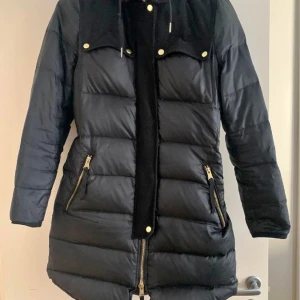 J.Lindeberg - Säljer min jacka från J Lindeberg.  storlek 38.  Och svart med guldiga detaljer och svarta ullpartier. Modellen heter Dilana lux och är 90% dun och 10% fjäder. Midjan går att justera de bak med knappar och luvan är avtagbar.