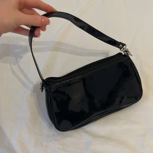 Stilren handväska svart  - Svart blank väska med silvriga detaljer och dragkedja. Inga fickor i. Oanvänd. Enkel att styla. 💕