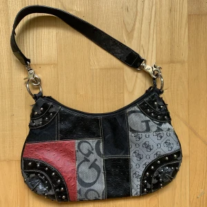 Väska - Guess inspirerad väska med röda, gråa och svarta detaljer med nitar. Den har några skavanker och tappar lite av utsidan därav priset