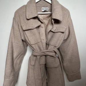 Kappa  - Ljusbeige kappa från NA-KD med skärp i midjan. Superfin färg och längden är perfekt! Endast använd ett fåtal gånger. Storlek 38. Nypris 600kr