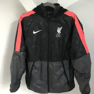 Tunn träningsjacka från Nike, fotbollsklubben Liverpool.  Vindtät, fodrad med tunt meshtyg. Fint skick. Luvan kan rullas ihop och stoppas in i kragen.