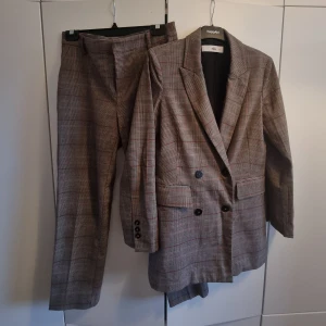 Rutigt brunt kostymset från Ellos - Rutigt kostymset med röda detaljer från Ellos. Cigarettformade kostymbyxor med fickor och hällor. Dubbelknäppt kavaj med fickor. Storlek 36 i kavaj, storlek 38 i byxor.