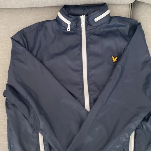 Lyle & scott vindjacka. - En marinblå Lyle & scott jacka med tunt material. Jackan har även en inbyggd luva som man kan ta fram ifall man vill det. Jackan har inga skador och är k bra skick. Storleken är i large. Ordinarie pris är 899 kronor och säljs pga fel storlek.