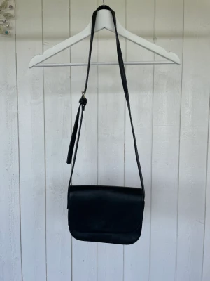 Axelremsväska  - En enkel svart liten axelremsväska med reglerbart band.  Innerfickans söm är lite trasig. 