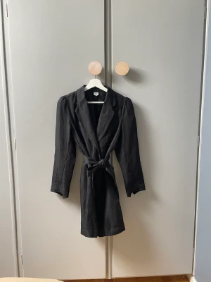 Linne kavaj/klänning med knytband i midjan och knappar strl 34 (36)  - Fin linnekavaj som passar som en klänning. Ett knytband i midjan och knappar längst med framsidan samt en ficka på vänstra yttersidan. Fina detaljer längst ner vid ärmarna. Små slitage, mycket sparsamt använd. I strl 34 men passar 36a. 