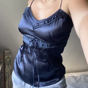 Blå topp i satin  - Säljer en jättefin mörkblå topp i ett satin liknande material. Linnet har en rosett längst fram, räfflor i tyget, samt spetsdetaljer. 
