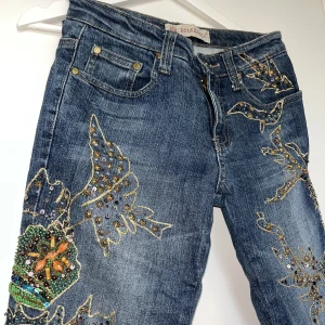Jeans med stenar - Blå jeans med strass, guldtråd, pärlor, rhinestones och paljetter från Rosa & Rosa. Utsvängda och lågmidjade. Fint skick. Innerbenslängd ca 72cm, Grenhöjd ca 21cm, midjemått ca 76cm. Jag är S och 175cm lång och de är för små på mig. 