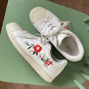 Vita sneakers med fin detalj - Snygga vita sneakers strl 41 med broderade blommor på sidan. Väldigt lite använda, men trots allt slitna så kvalitén är inte den bästa, därav pris.