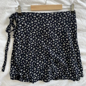 Brandy Melville omlott kjol med blommigt mönster - En omlottkjol med blommigt mönster från brandy Melville. Kjolen är mörkblå med krämvita blommor. Den är helt justerbar i banden, så passar därav flera storlekar. Sparsamt använd, jättefint skick. Köpt i Hawaii 💗