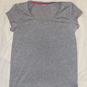 T-Shirt - Grå t-shirt med rosa sömmar och ett rosa fjärilstryck vid nacken. T-Shirten är från Me&I i storlek M.