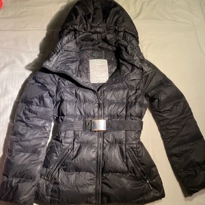 Svart dunjacka  - Storlek 36 och märket är Beneton. Detaljer i silver samt ett tillhörande midjeband som är enkelt att plocka av om så önskas. Avtagbar luva. Finns ett knappt synbart hål i högra hörnet. 