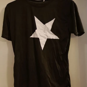 Svart T-shirt med stjärna  - Svart T-shirt med tryck på framsidan. Nåt slags polyester-material, är lite genomskinlig. Aldrig använd.