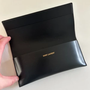 Saint Laurent glasögonfodral/clutch - Oanvänt svart läderfodral av märke Saint Laurent. 19 cm långt, kan användas som liten clutch eller som glasögonfodral. Instansad logo på framsidan och tryckt guldlogo på insidan.
