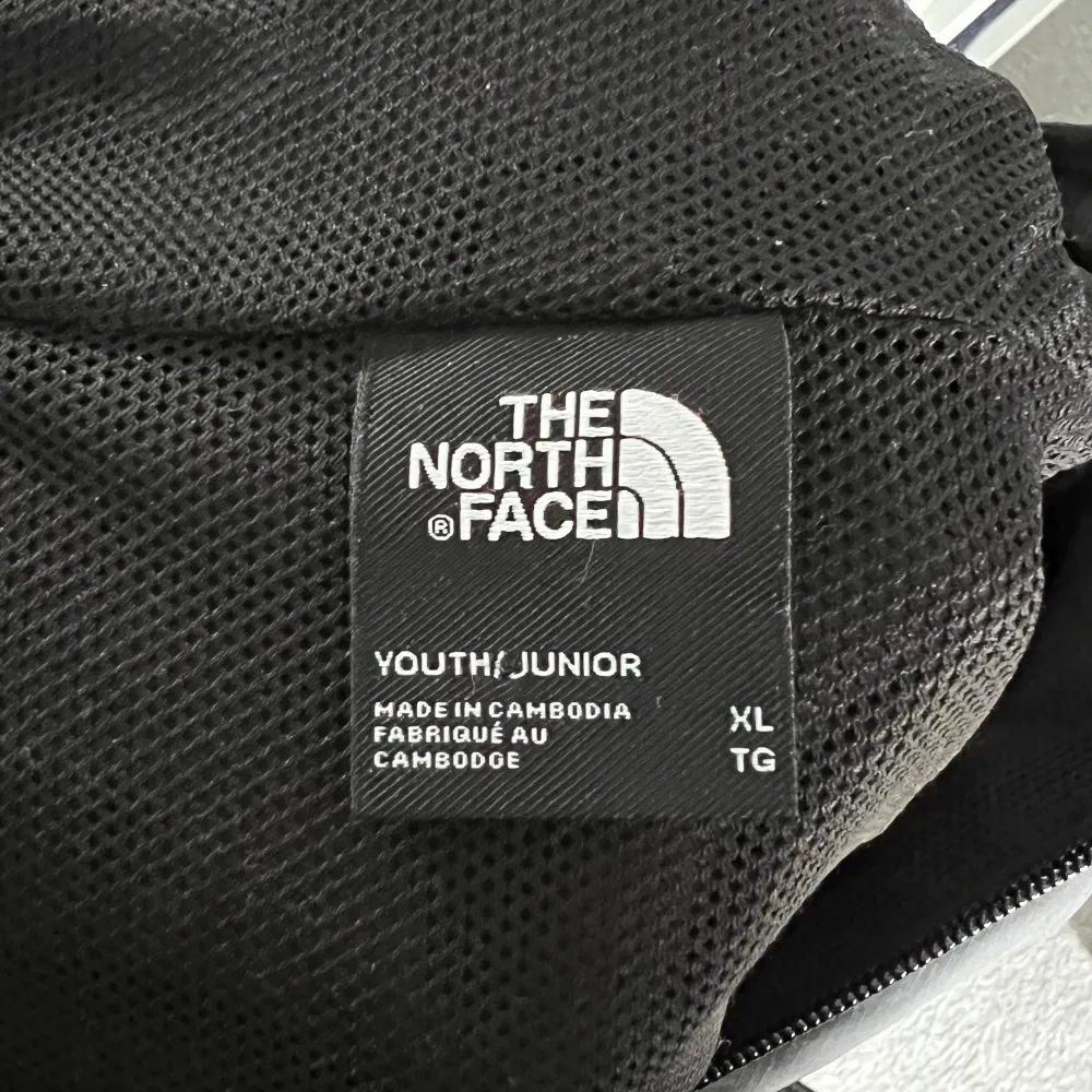 Vindjacka med huva från The North Face, svart/grå i storlek Youth XL men liknar även XS-S herr/dam. Färgskillnaderna mellan svart och grått inte lika skarpa som på bild. Använd ett par tillfällen förra året innan den blev för liten. . Jackor.