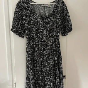 Svart blommig klänning - En svart klänning med vita blommor och korta ärmar. Klänningen har en knäppning framtill och snörning i ryggen. Den är kort i längden och har en rund halsringning.