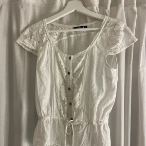 Vit topp från Lindex - En vit topp från Lindex med spetsdetaljer på ärmarna och längs med knäppningen framtill. toppen har korta ärmar och en knytning i midjan för en justerbar passform. Den är dekorerad med knappar framtill.