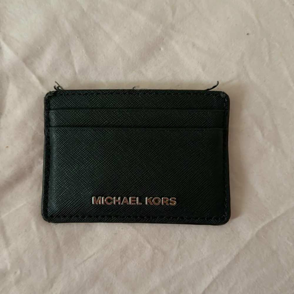 En elegant svart plånbok från Michael Kors med flera kortfack. Plånboken har en stilren design med Michael Kors-logotypen i guld på framsidan. Halvt sliten som ni ser med två tre lösa trådar men kan klippas bort. Väskor.