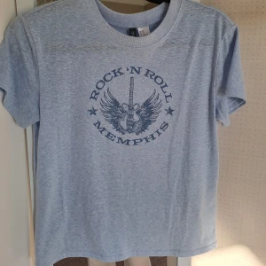 Ljusblå t-shirt med tryck - En ljusblå t-shirt med ett tryck av en gitarr med vingar och text. T-shirten har korta ärmar, en rund halsringning och väldigt skönt material.✨️