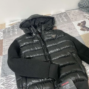 Svart jacka från Prada - En svart jacka från Prada med huva och dragkedja. Jackan har stickade ärmar och är vadderad med dun och vit ejderdun. Framsidan och huvan är gjorda av nylon medan ryggen och ärmarna är av ull. Jackan har en blank yta och en dragkedja framtill. Den har även tvà fickor pa sidorna och en innerficka pa jackan. Storleken är i storlek M men passar som S. Den är bara prövad men aldrig använd. Jackan värmer jätte bra och passar perfekt till kommande väder, priset är inte hugget i sten utan går att diskutera Sälj pågrund av den har aldrig kommit till användning.