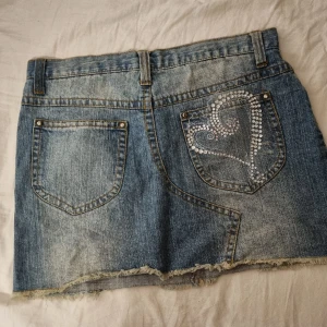 Unik jeanskjol med strass hjärta  - Unik jeanskjol med ett hjärta i strass på baksidan. Den yttre sömmen har gått up cirka 2cm på ett ställe, går enkelt att nästa för hand. 