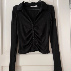 Topp NA-KD  - Svart topp från NA-KD i storlek S. Rynkad framtill och knäpps med knappar. Stretchigt och lite ribbat material. Liten defekt (se bild) men påverkar inte användningen och det syns knappt. Använd ett fåtal gånger, i bra skick annars. Finns fler bilder