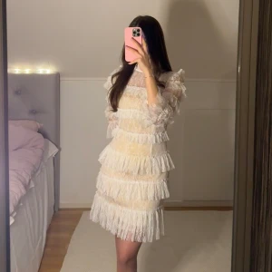 By Malina Carmine klänning - Carmine klänning i spets med hög krage, långa ärmar och kort kjol täckt i volanger. PERFEKT INFÖR STUDENTEN😍  DETALJER • Miniklänning med spets-volang design och en tight passform • Polokrage i spets • Dragkedja längs ryggen