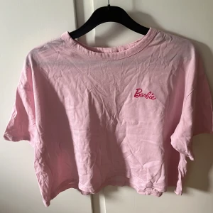 Rosa Barbie t-shirt - En rosa t-shirt med Barbie-logotypen tryckt på bröstet. T-shirten är kortärmad och har en croppad design.