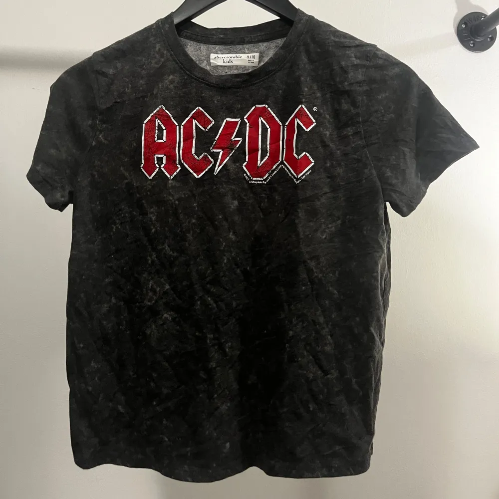 En svart t-shirt från Abercrombie Kids med ett stort rött och vitt AC/DC-tryck på framsidan. T-shirten har korta ärmar och en rund halsringning.. T-shirts.