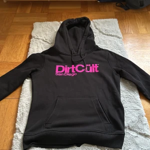 Dirtcült hoodie  - Dirtcült hoodie med rosa text säljes.  Den är köpt för cirka 2-3 år sedan men är knappt använd.  Storlek L, väldigt små i storlekarna. Jag har annars S