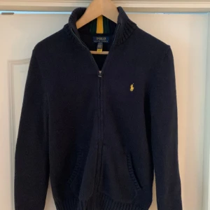 Ralph Lauren tröja full zip - Helt ny mörkblå stickad Polo Ralph Lauren tröja med dragkedja och fickor.   Nypris 1395 kr.  Inköpt på NK, kvitto finns.  Endast provad men lapparna borttagna så jag kan inte lämna tillbaka den.  Storleken i tröjan är 14-16 (L) motsvarar ung XS i vuxenstl