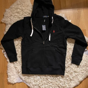 Polo Ralph Lauren Zip-Hoodie - Presenting a sleek black Polo Ralph Lauren zip-up cardigan, in impeccable 10/10 condition and retaining its pristine price tag. This sophisticated piece not only exudes a sense of timeless style but also showcases the brand's commitment to quality. 