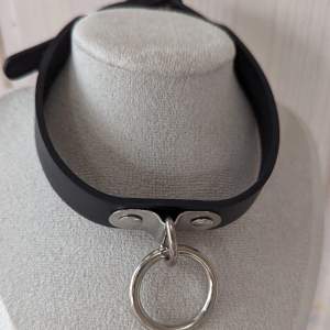 Gotiskt choker-halsband från Restyle.Har en justerbar krage. Dekorerad med en stor metall-O-ring.  Ett perfekt smycke till en riktigt gotisk look! Material 90% polyuretan och 10% metall 