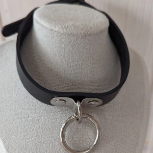 Gotisk choker-halsband - Gotiskt choker-halsband från Restyle.Har en justerbar krage. Dekorerad med en stor metall-O-ring.  Ett perfekt smycke till en riktigt gotisk look! Material 90% polyuretan och 10% metall 