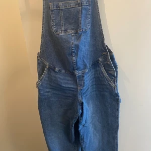 MAMA Ankellång hängselbyxa i denim. Storlek M. - Ett par ankellånga hängselbyxor i stadig bomullsdenim.  Byxorna är bara provat. Reglerbara hängslen, knappar i ena sidan och fuskgylf. Bröstficka, snedställda sidfickor och bakfickor. Lätt avsmalnande ben med hammarslinga och en benficka. Insida ben: