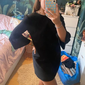 Svart klänning med ärmar - Ganska tjockt tyg så håller värmen bra. Fina 3/4 ärmar och volanger i slutet. Mycket skön. Lagom längd. 