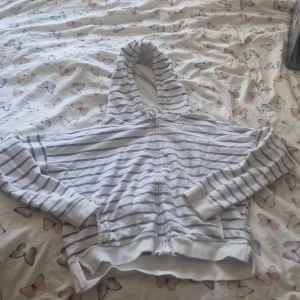 En vit blårandig tjocktröja  - En skön tjocktröja som är vit och har blåa ränder. Den har dragkedja, luva och snören så man kan välja hur tajt luvan ska sitta. ❤️