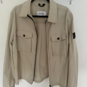 Overshirt - Snygg overshirt från Calvin Klein, aldrig använd utan bara prövad. Ljusbeige med svarta detaljer och dragkedja. Se sista bilderna för hur den sitter på. Färgen är ljusare än på bild. Storlek L. Köpt för 1700kr. Priset går att sänkas vid snabb affär:)