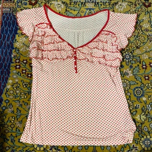 Prickig topp - Jättefin vit topp med röda prickar, fina detaljer som volanger och knappar. Fint skick. Finns extraknappar på insidan ifall någon skulle trilla av.