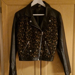 Kort jacka i fejkskinn - Snygg, kort jacka i fejkskinn och med en framsida i leopard av någon form av fleecetyg. Två fickor fram. Knappt använd, skick 9/10. Strl 38.  Hund finns i hemmet. 