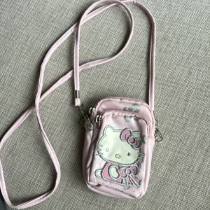 Mobilväska Hello Kitty - Söt liten väska för mobilen och små saker man vill ha med sig, t ex läppglans, kort och liknande. Lite sliten på framsidan, se bild, annars i fint skick. Storlek ca 14x10 cm. Tre fack som stängs med dragkedja