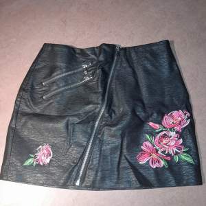 En svart kjol i skinnimitation med dragkedja samt ros-broderier framtill. Den är i storlek 36 från H&M, helt ny, köpt för 249 kr. Måtten på den är: 35cm lång, 36cm i midjan och 47 cm längst ner. 