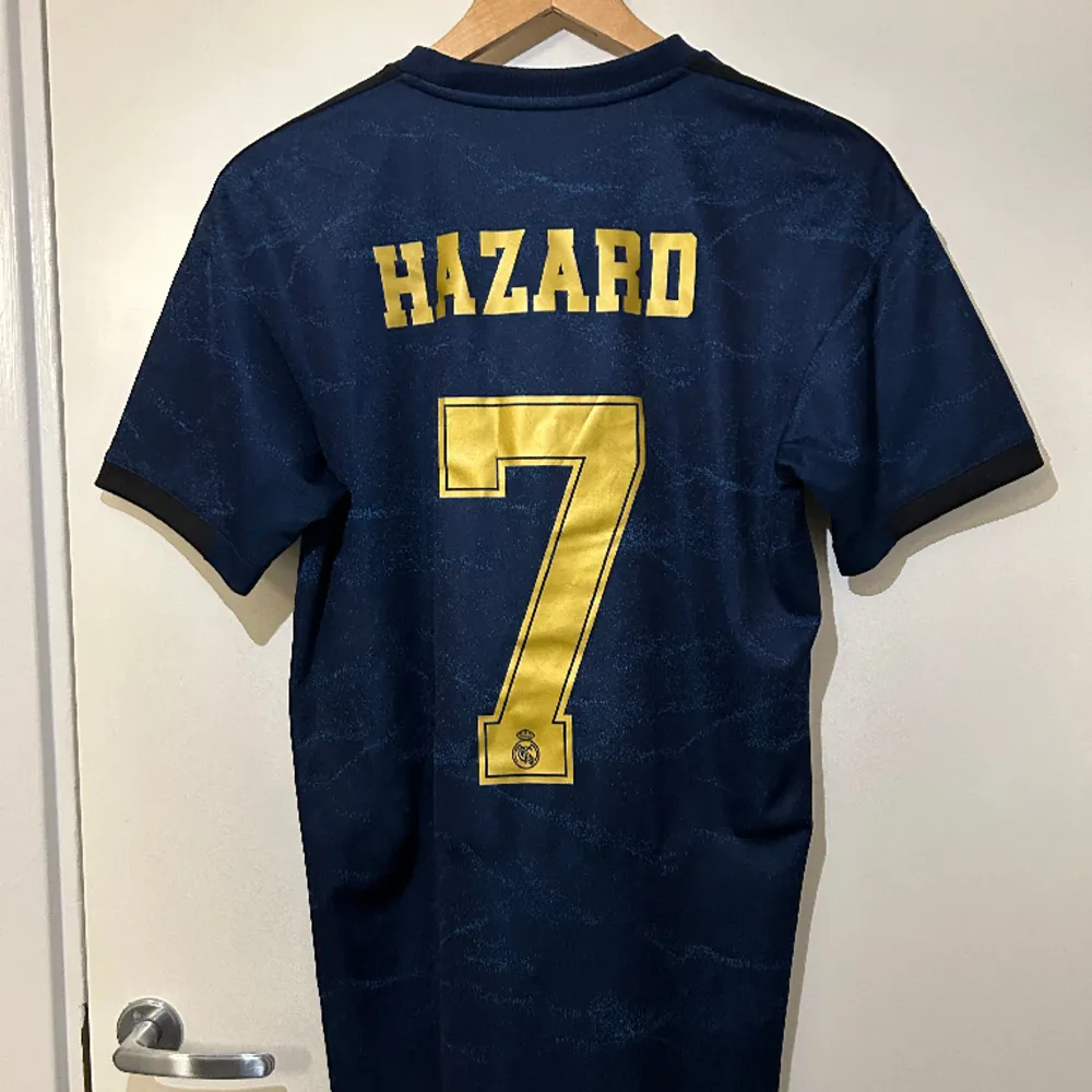 Äkta mörkblå Real Madrid 2019/20 borta fotbollströja med Hazard och nummer 7 på ryggen. Guldig text och detaljer samt svarta ränder på ärmarna i storlek S. Använd få gånger. . T-shirts.